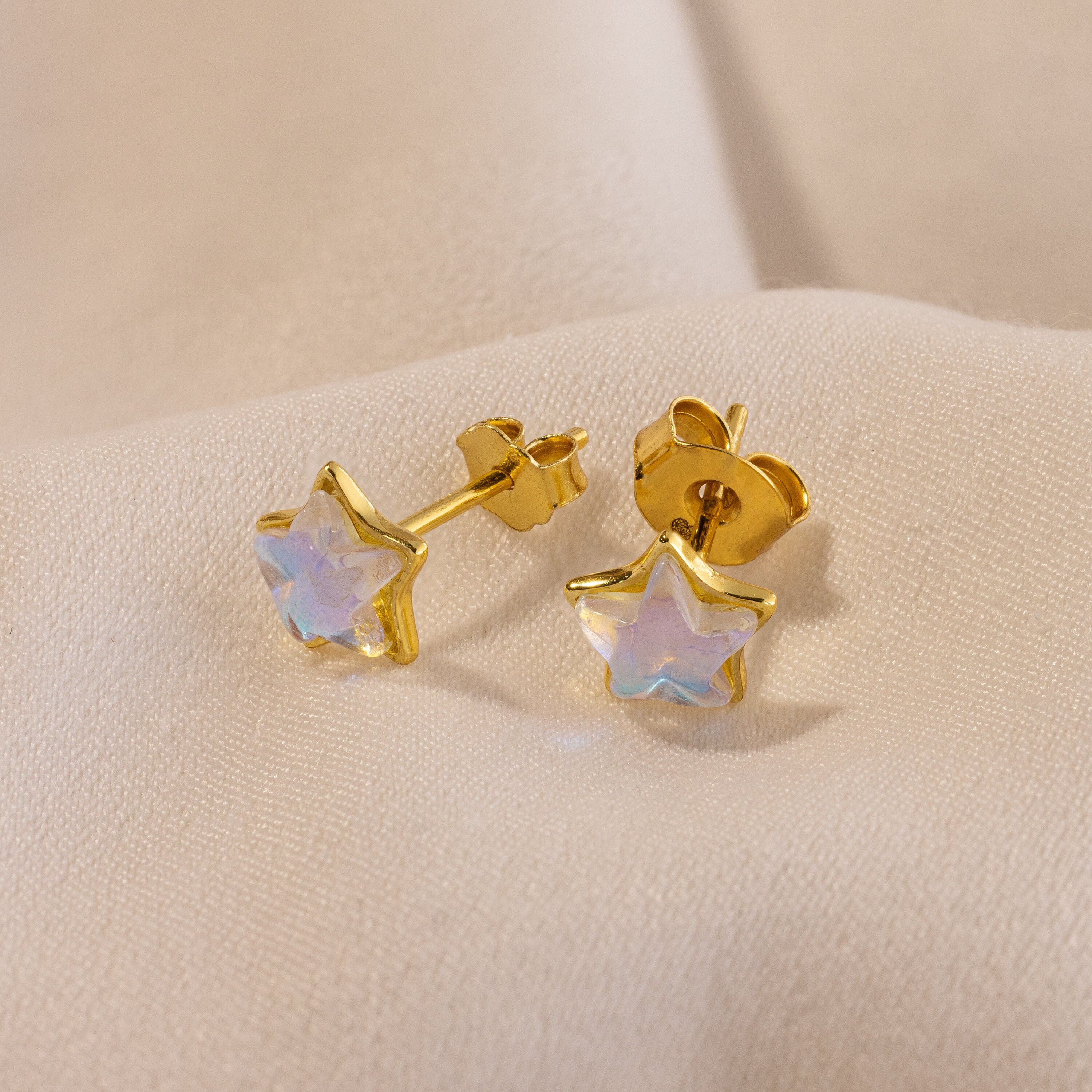 Moonstone Star Studs with gold star-shaped design and iridescent centers shimmer on a soft beige fabric background.