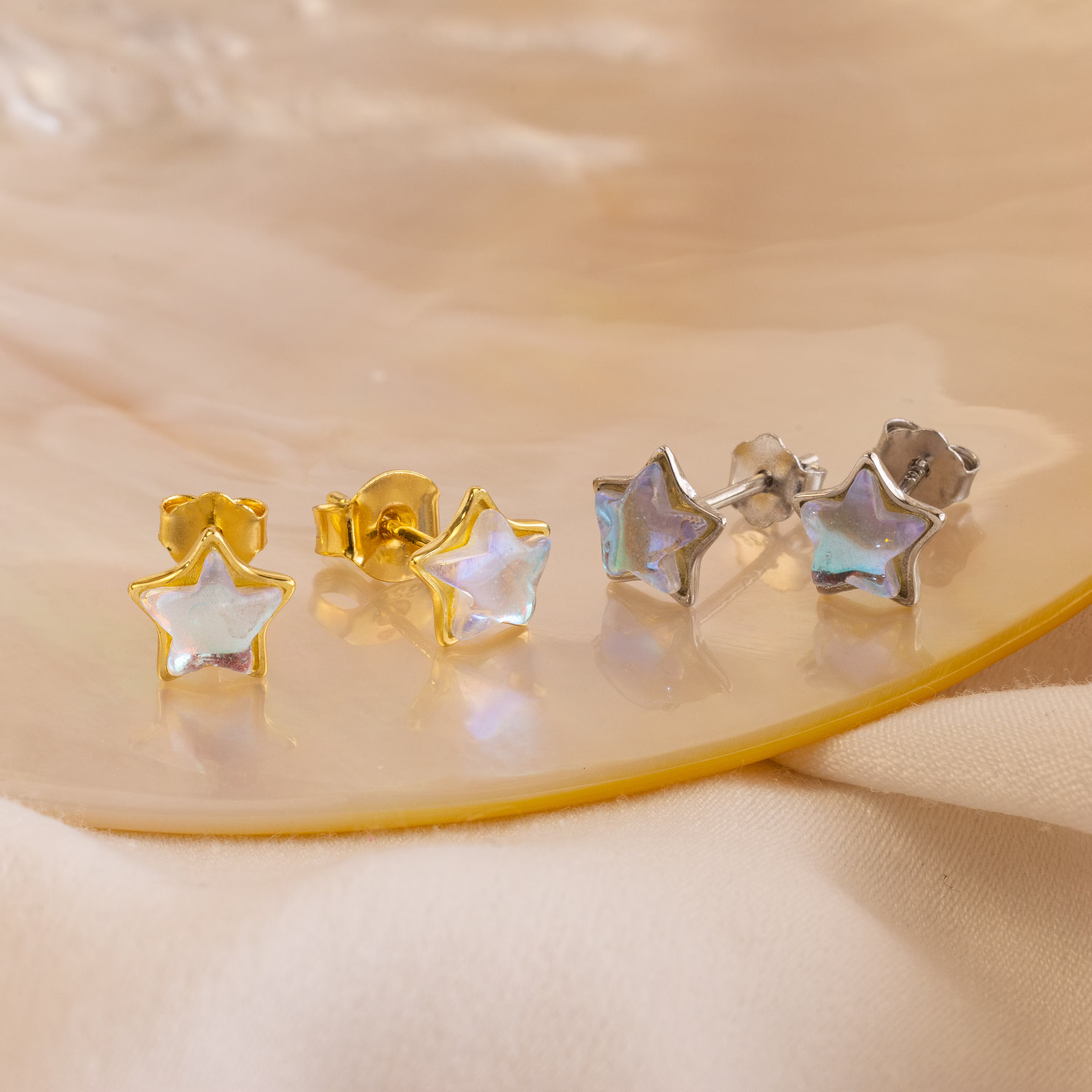 Two pairs of Moonstone Star Studs—star-shaped earrings in gold and silver—are displayed on a beige surface.