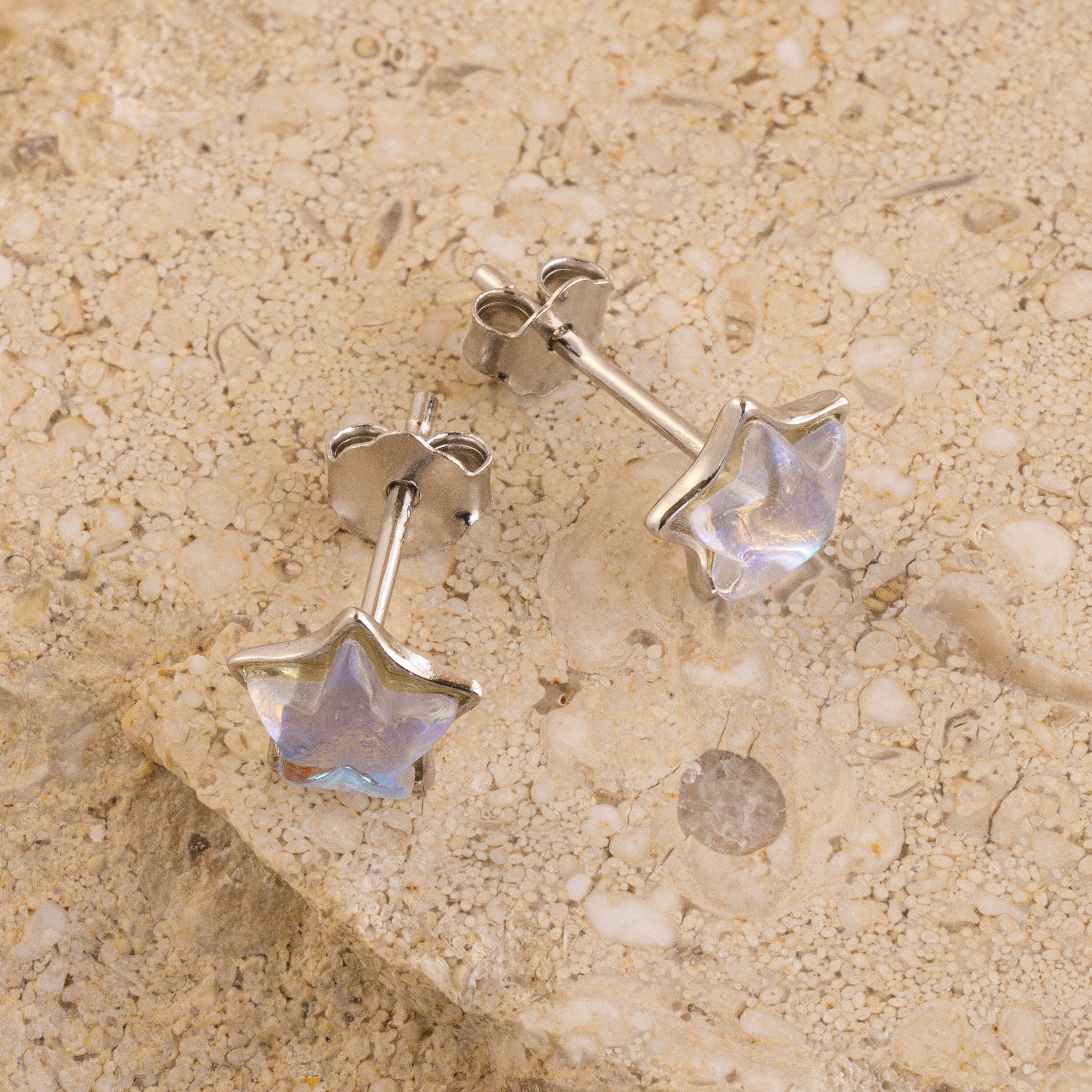 Moonstone Star Studs are silver star-shaped earrings featuring iridescent centers, radiating celestial charm and shown on a textured beige stone surface.