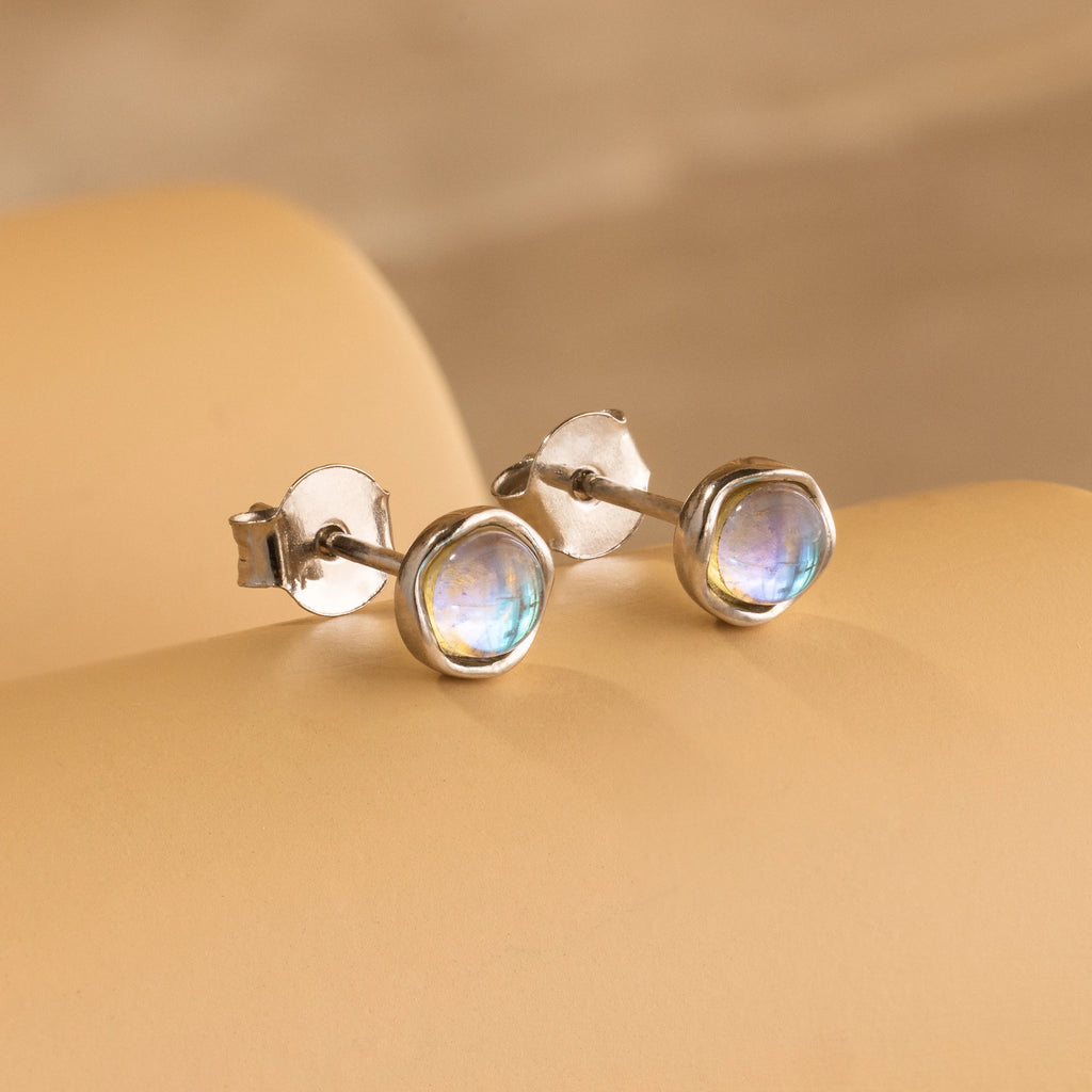 Experience celestial beauty with the Moonstone Studs—silver earrings featuring round, iridescent moonstones, elegantly displayed on a soft beige surface.