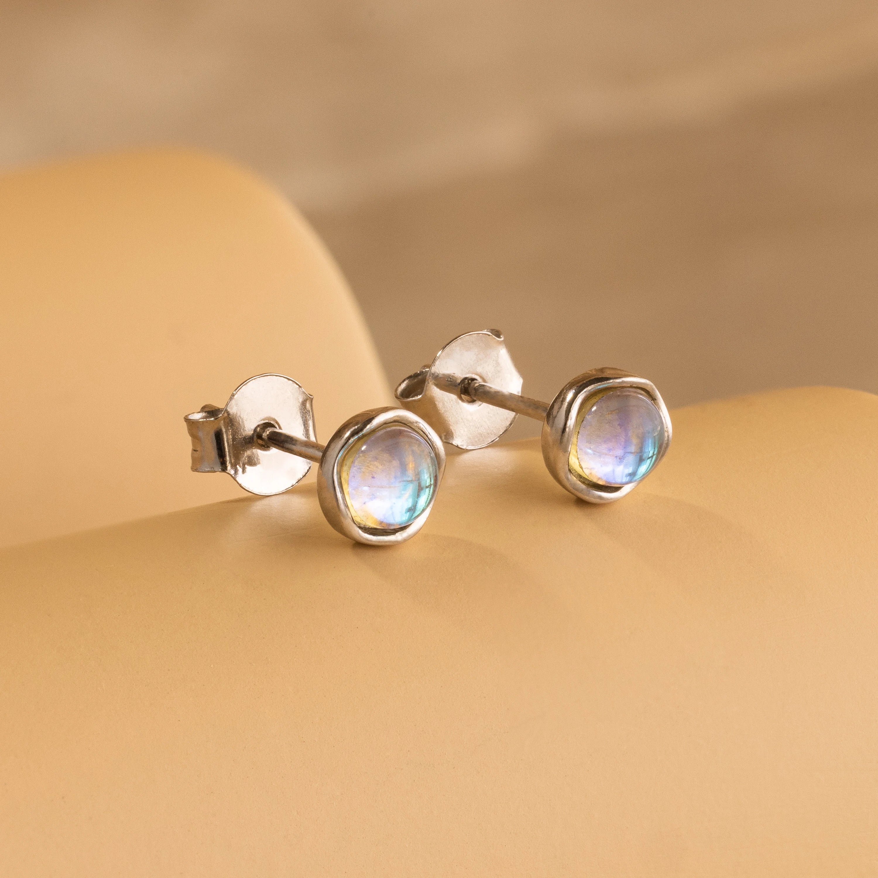 Experience celestial beauty with the Moonstone Studs—silver earrings featuring round, iridescent moonstones, elegantly displayed on a soft beige surface.
