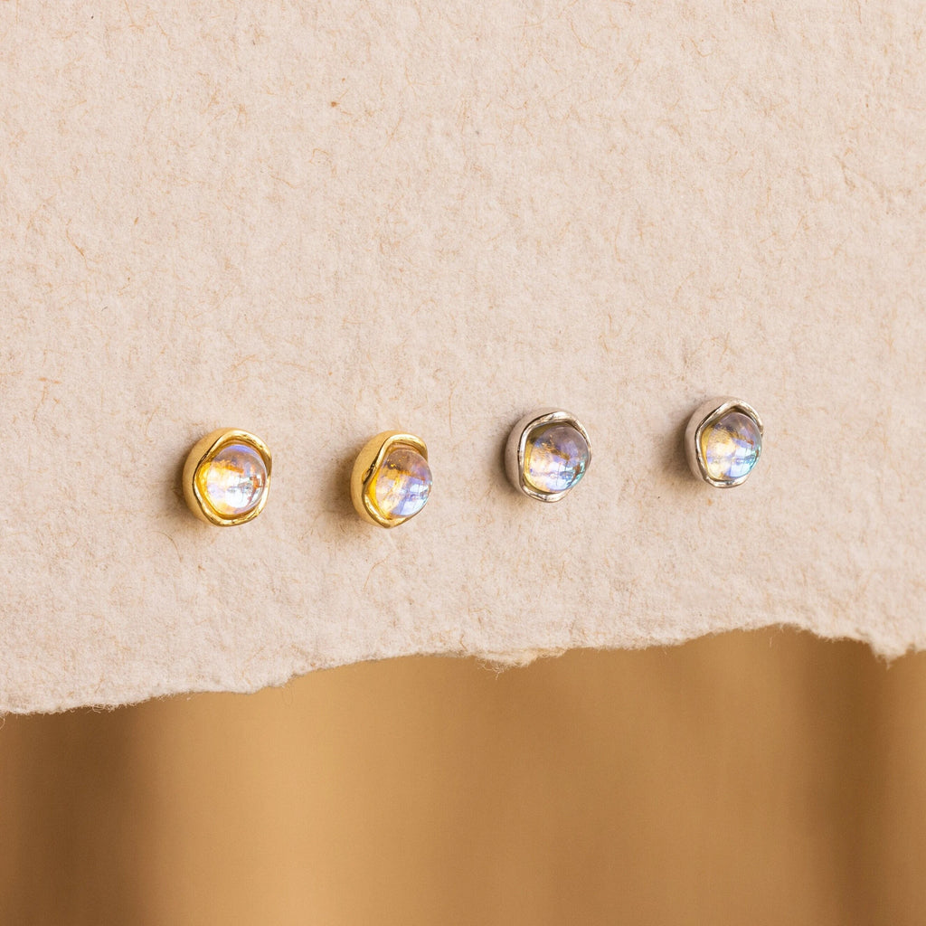 Four small round Moonstone Studs—two in gold and two in silver settings—are displayed on textured beige paper, perfect for fans of delicate celestial jewelry.
