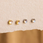 Four small round Moonstone Studs—two in gold and two in silver settings—are displayed on textured beige paper, perfect for fans of delicate celestial jewelry.