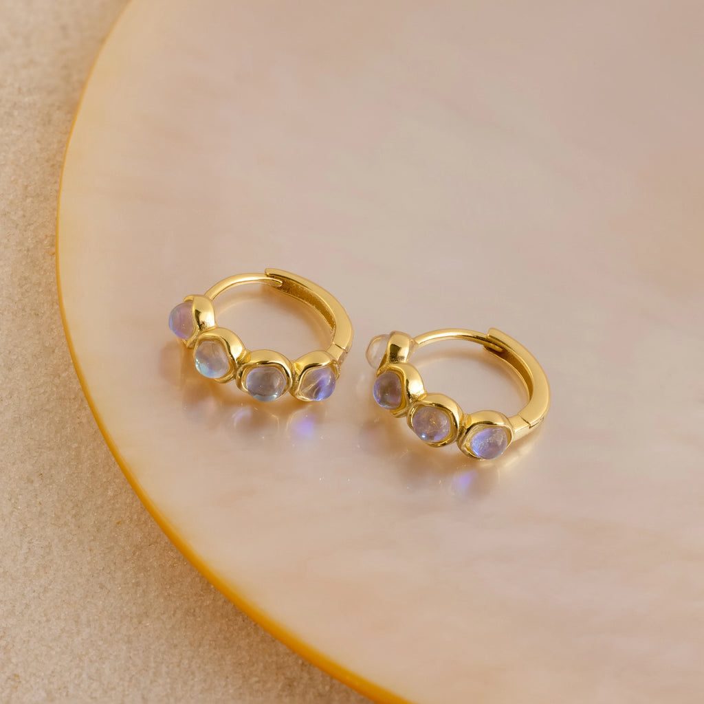 The Clear Moonstone Huggies are gold hoop earrings set with four round iridescent moonstone gems, elegantly displayed on a pale round ceramic plate.