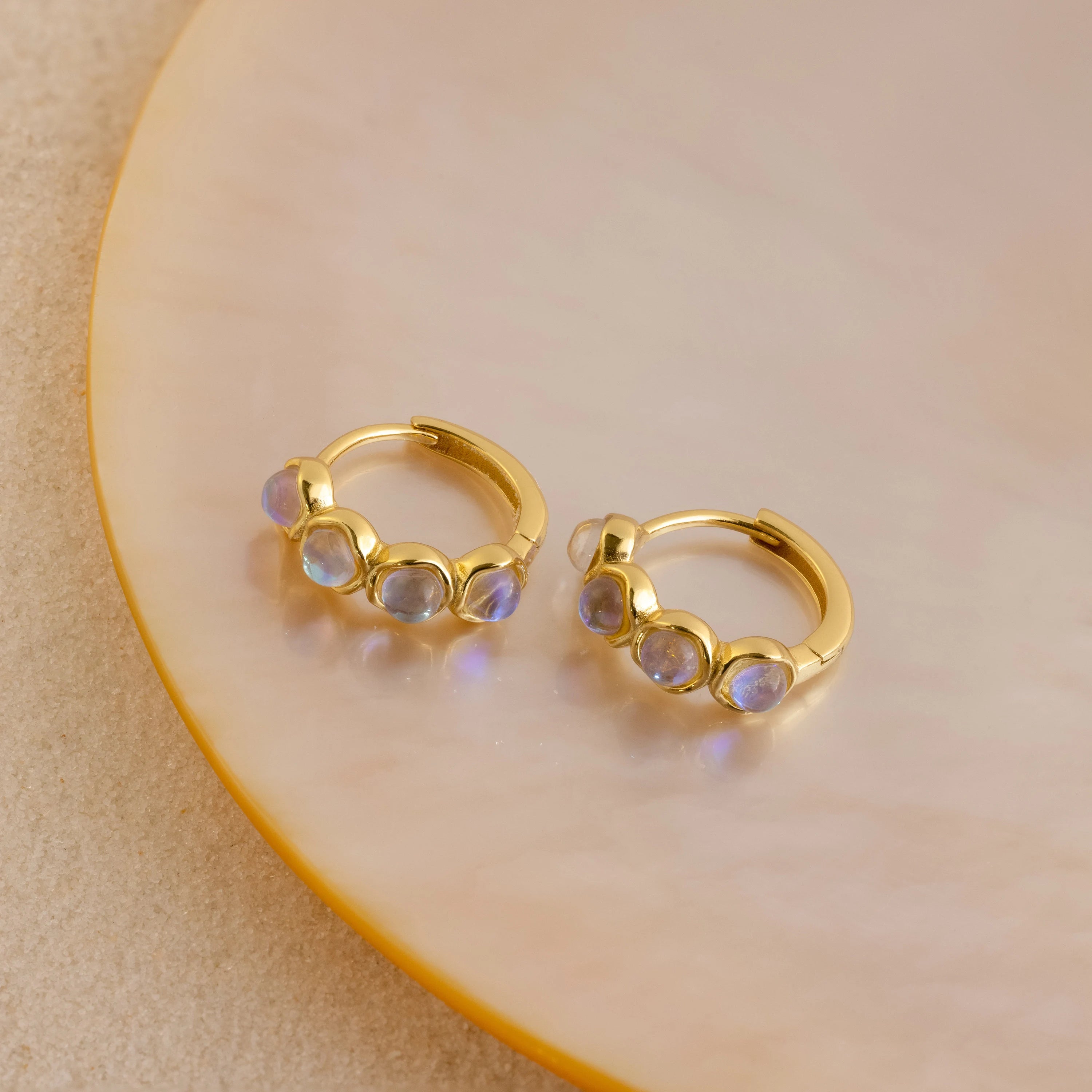 The Clear Moonstone Huggies are gold hoop earrings set with four round iridescent moonstone gems, elegantly displayed on a pale round ceramic plate.