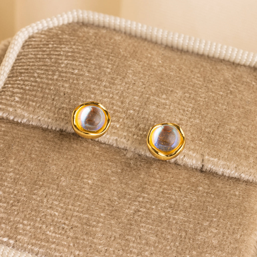 Moonstone Studs: Gold-rimmed, round earrings with clear stones, shown on a beige fabric display—ideal for celestial jewelry fans.