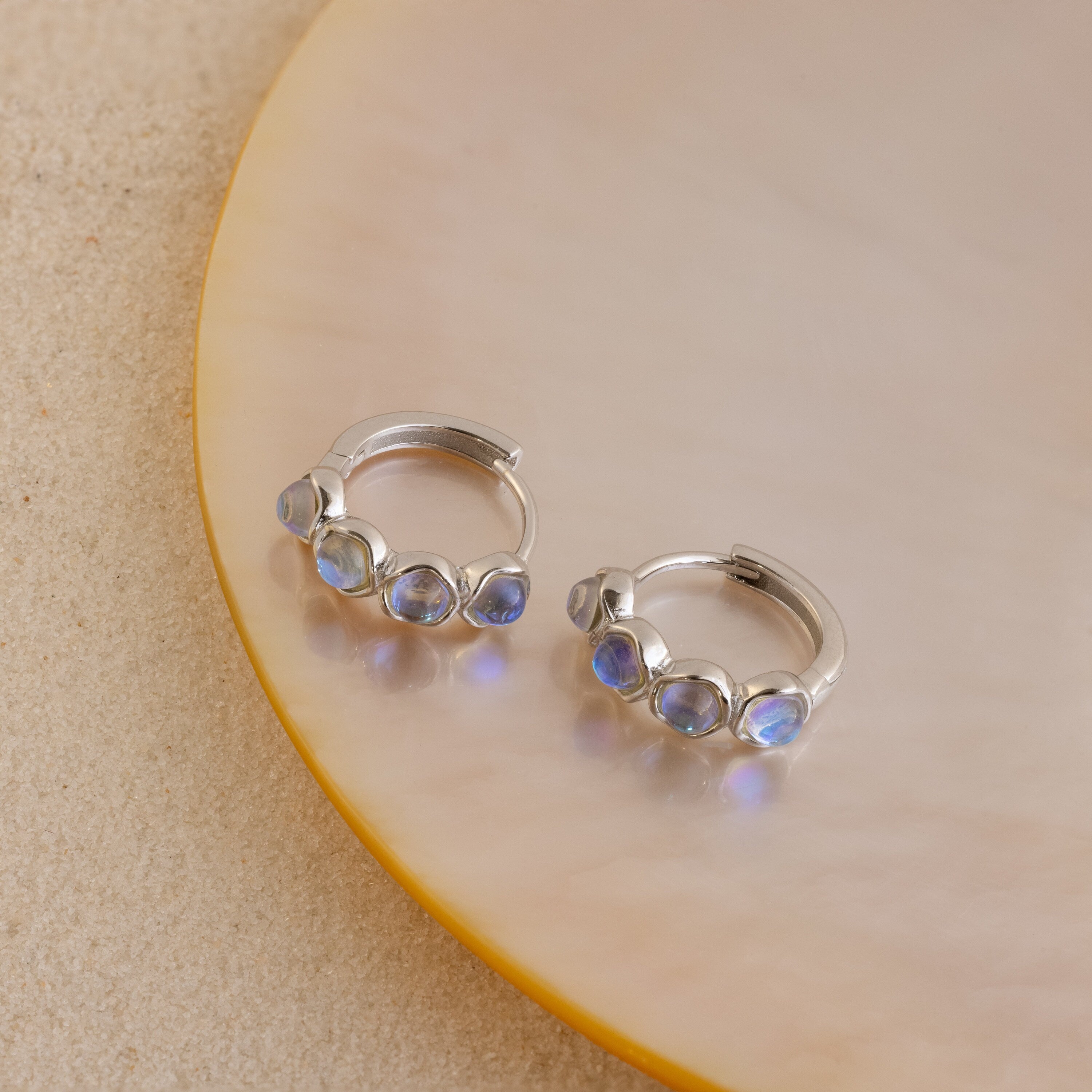 The Clear Moonstone Huggies feature elegant silver hoops set with four blue moonstones, showcased on a round cream and gold display.