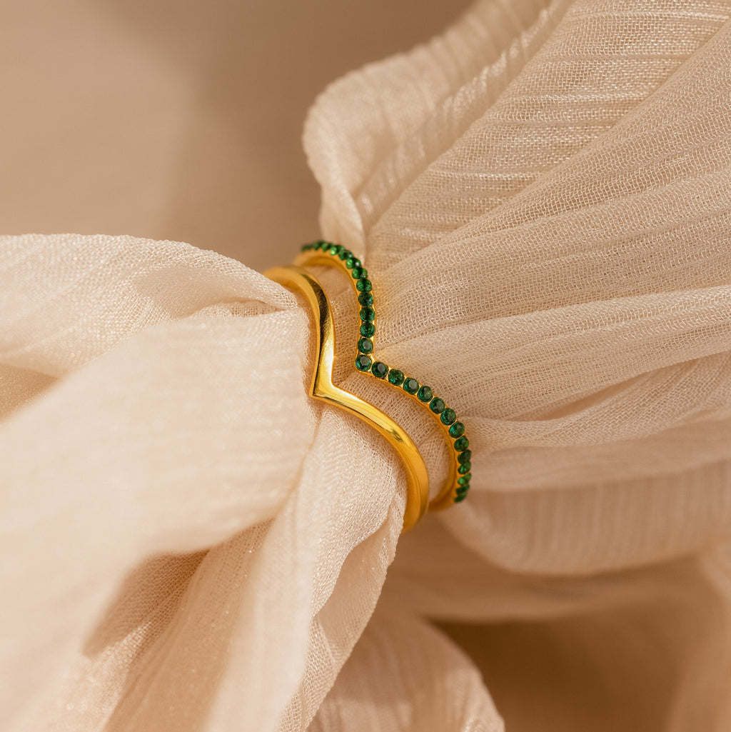 The Chevron Birthstone Rings Set, featuring gold and green bands, is displayed on beige textured fabric in a loose knot to showcase their distinctive chevron design and colorful birthstone accents.