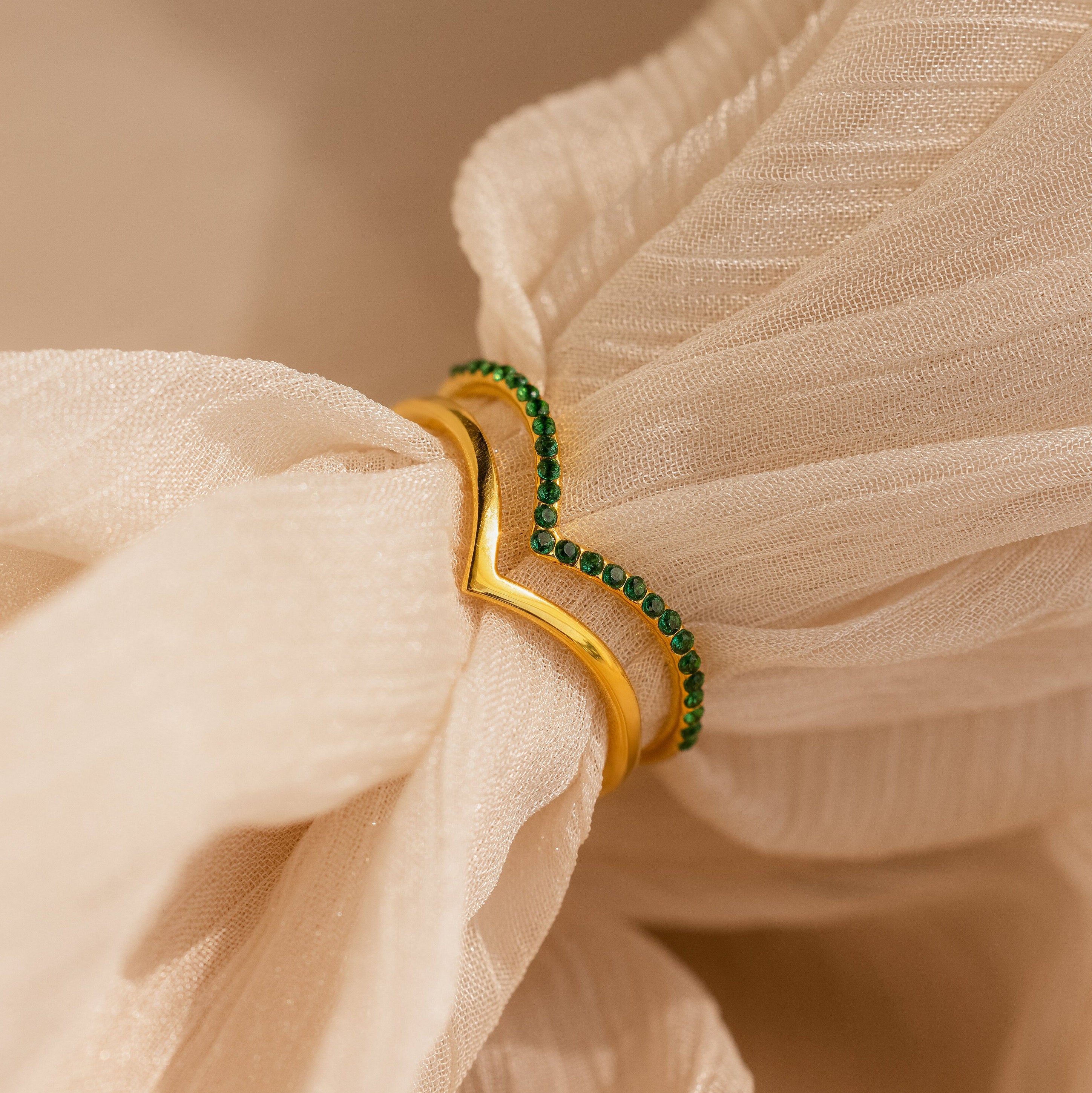 The Chevron Birthstone Rings Set, featuring gold and green bands, is displayed on beige textured fabric in a loose knot to showcase their distinctive chevron design and colorful birthstone accents.