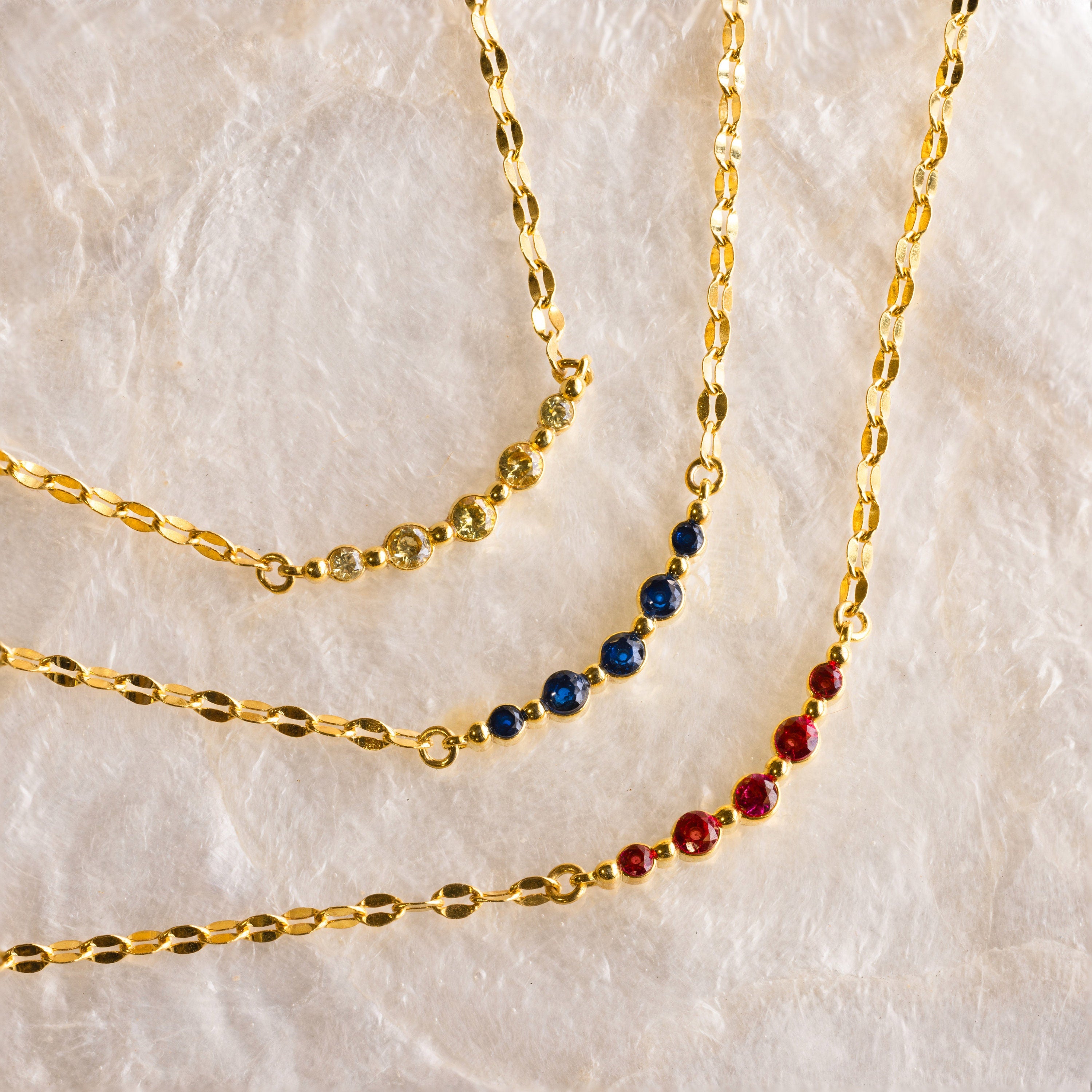 The Lace Birthstone Necklace features three gold chains with yellow, blue, and red gemstone beads on a neutral textured surface—ideal for a personalized, dainty birthstone gift.