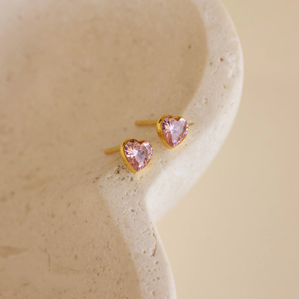 Bezel Heart Birthstone Studs feature pink heart-shaped gemstones and rest on a textured cream surface, highlighting the charming elegance of these stud earrings.