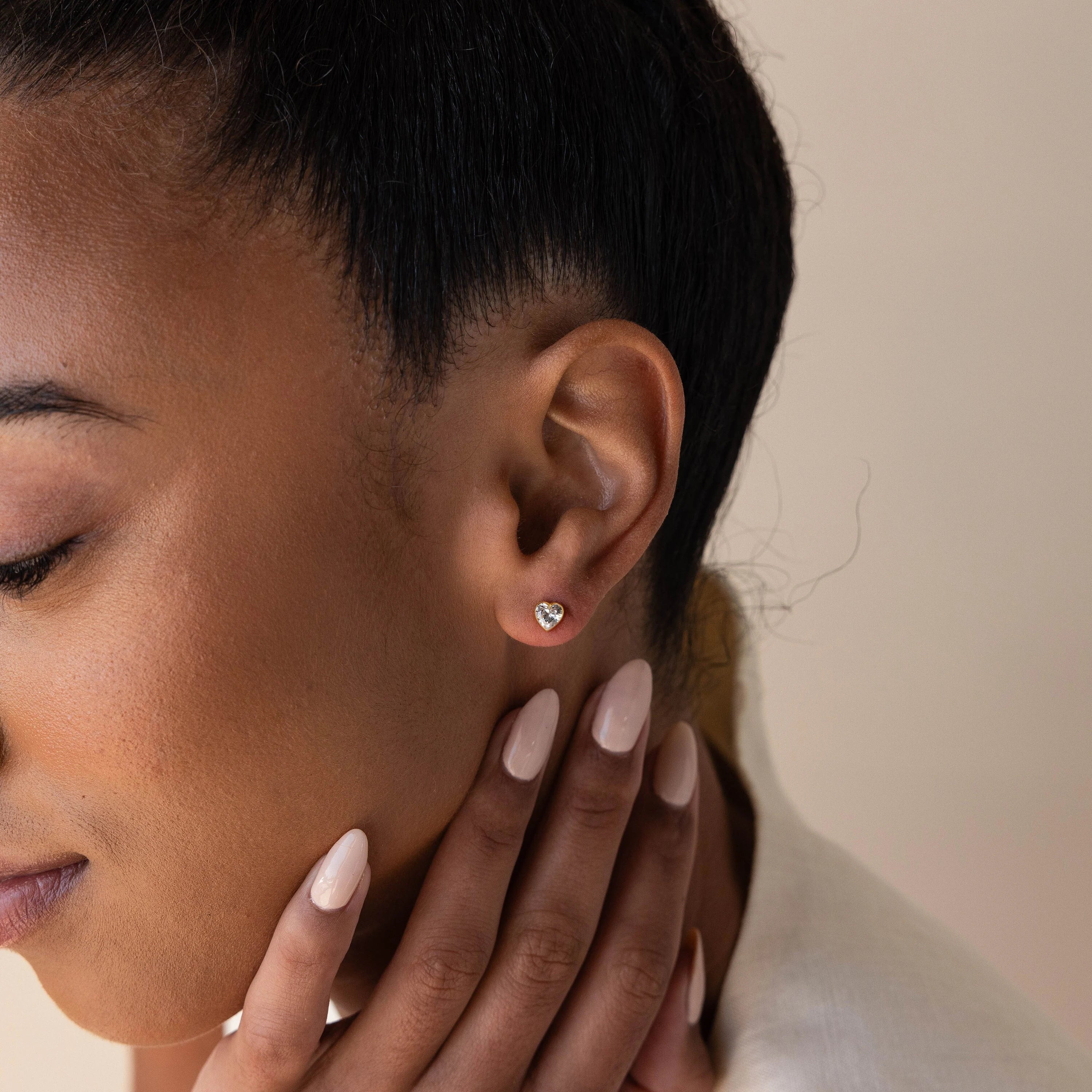 A woman touches her face, showcasing manicured nails and a Bezel Heart Birthstone Stud earring.