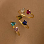 Six Duo Heart Birthstone Rings with heart-shaped gemstones are arranged in a spiral on a beige background, each resembling a personalized charm for a unique touch.