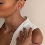 A woman wearing Bezel Heart Birthstone Studs and a blue heart-shaped gemstone ring holds her hand near her face.