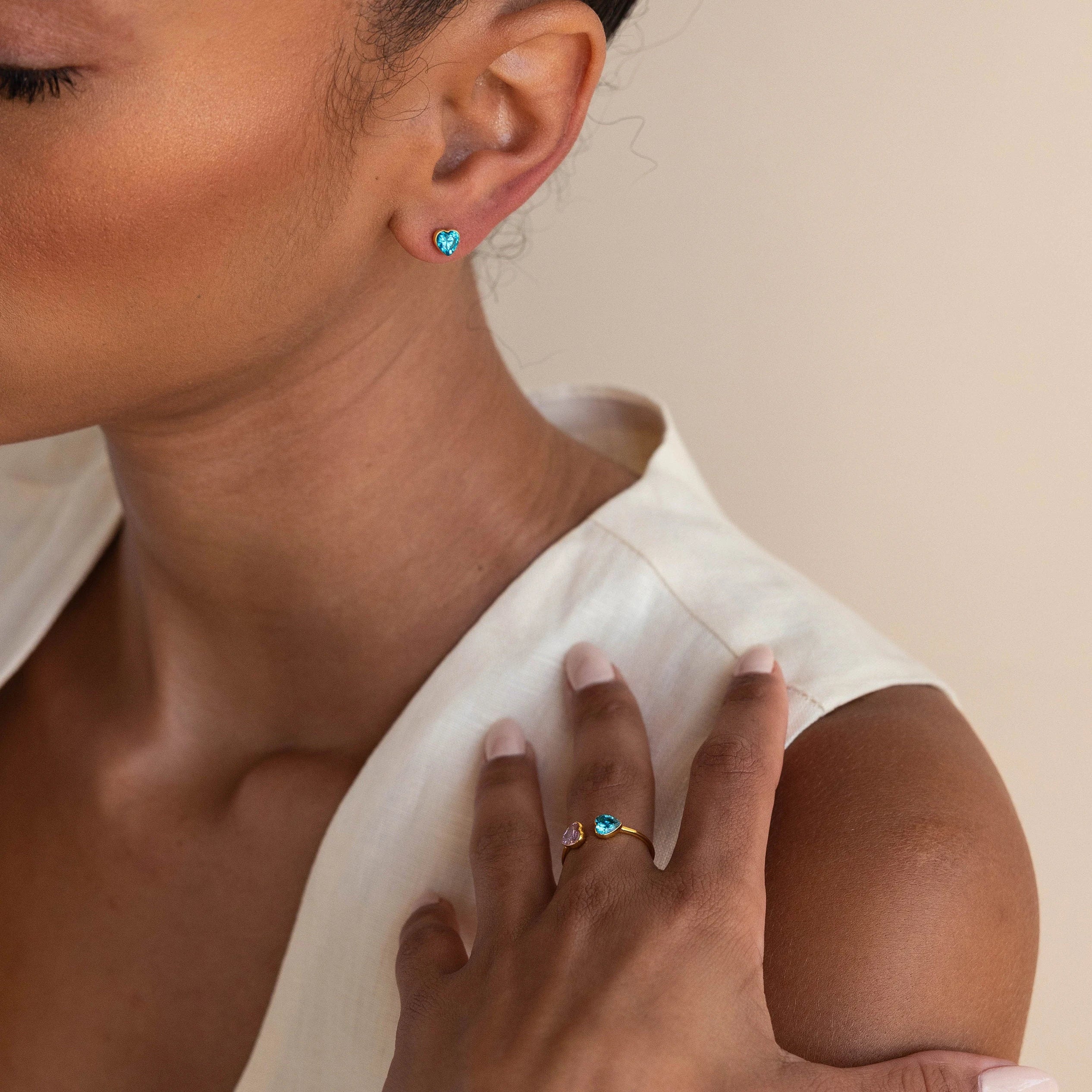 A woman wearing Bezel Heart Birthstone Studs and a blue heart-shaped gemstone ring holds her hand near her face.