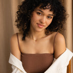 A woman with curly hair, in a brown top and white shirt, smiles softly in front of a beige curtain, her look completed by a delicate Tapered Baguette Birthstone Necklace.