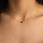 The Tapered Baguette Birthstone Necklace features a gold mirror chain and a small red rectangular pendant, creating an elegant accessory for any woman.