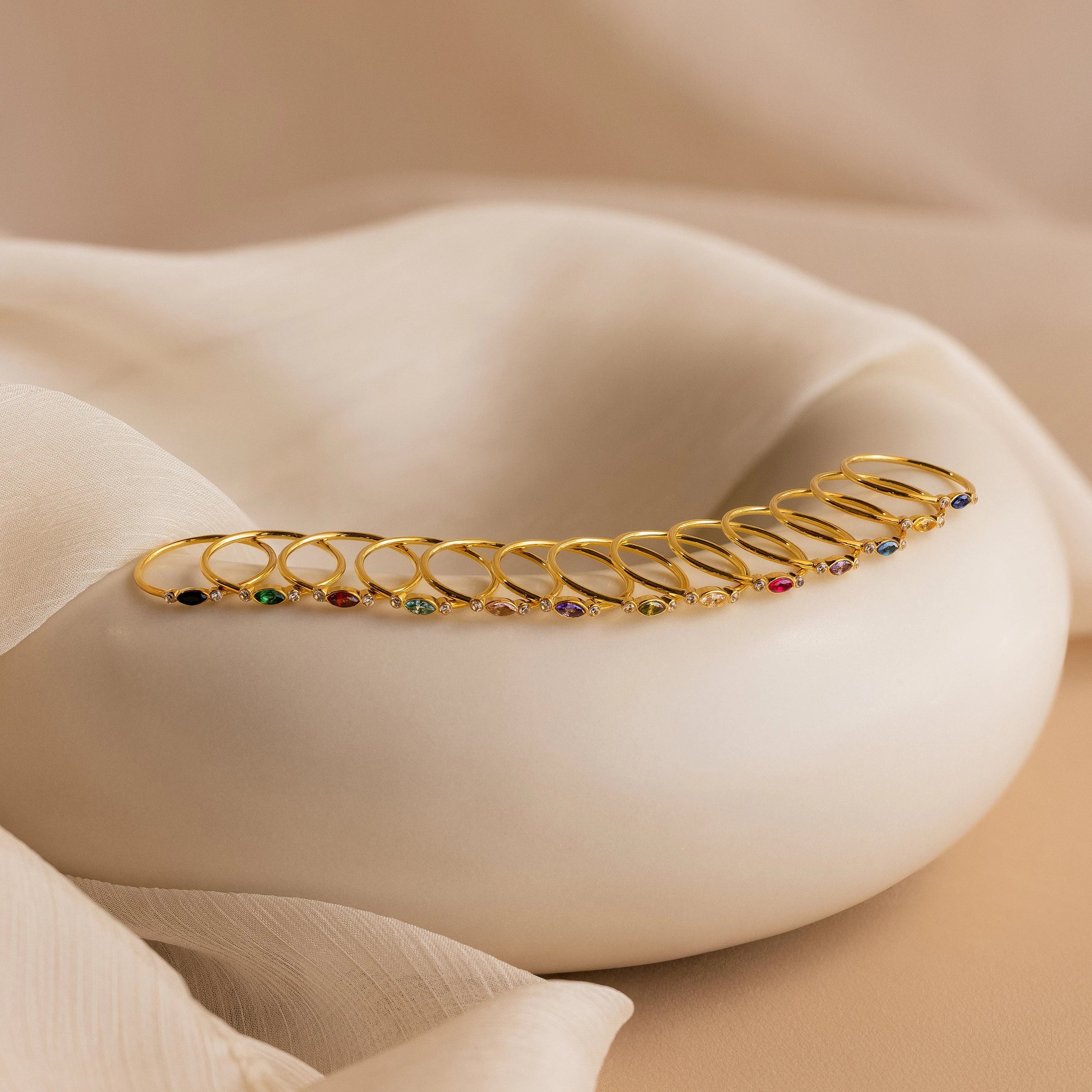 A row of gold rings, including the Marquise Bezel Birthstone Ring with colorful gemstones, displayed on a smooth white curved surface with fabric drapes.