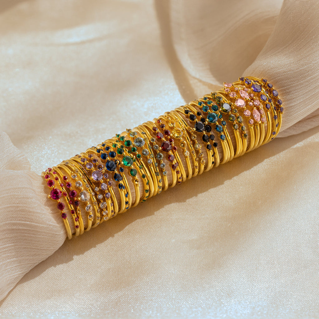 The Lace Birthstone Rings Set features gold bangles with colorful gemstones on soft, beige fabric—ideal for pairing with minimalist rings for a stylish, coordinated look.