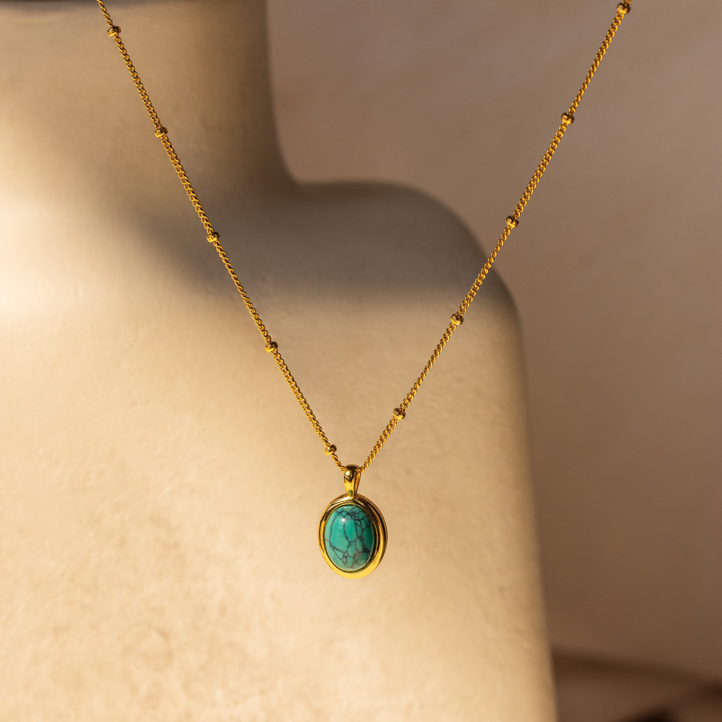 The Turquoise Pendant Necklace features a gold boho-chic chain with an oval turquoise pendant, shown on a neutral-toned mannequin bust.
