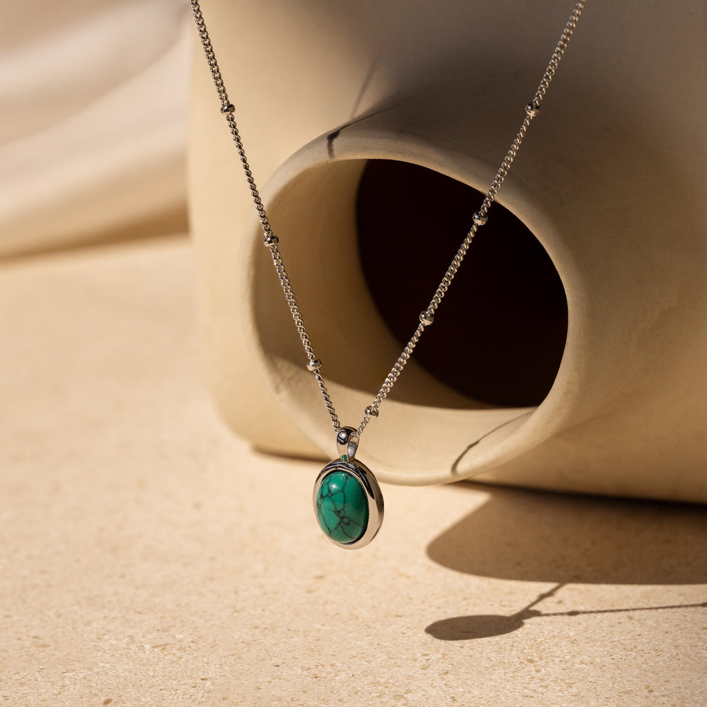 The Turquoise Pendant Necklace, featuring a silver boho-chic design with an oval turquoise pendant, is draped over a cream abstract ceramic vase.