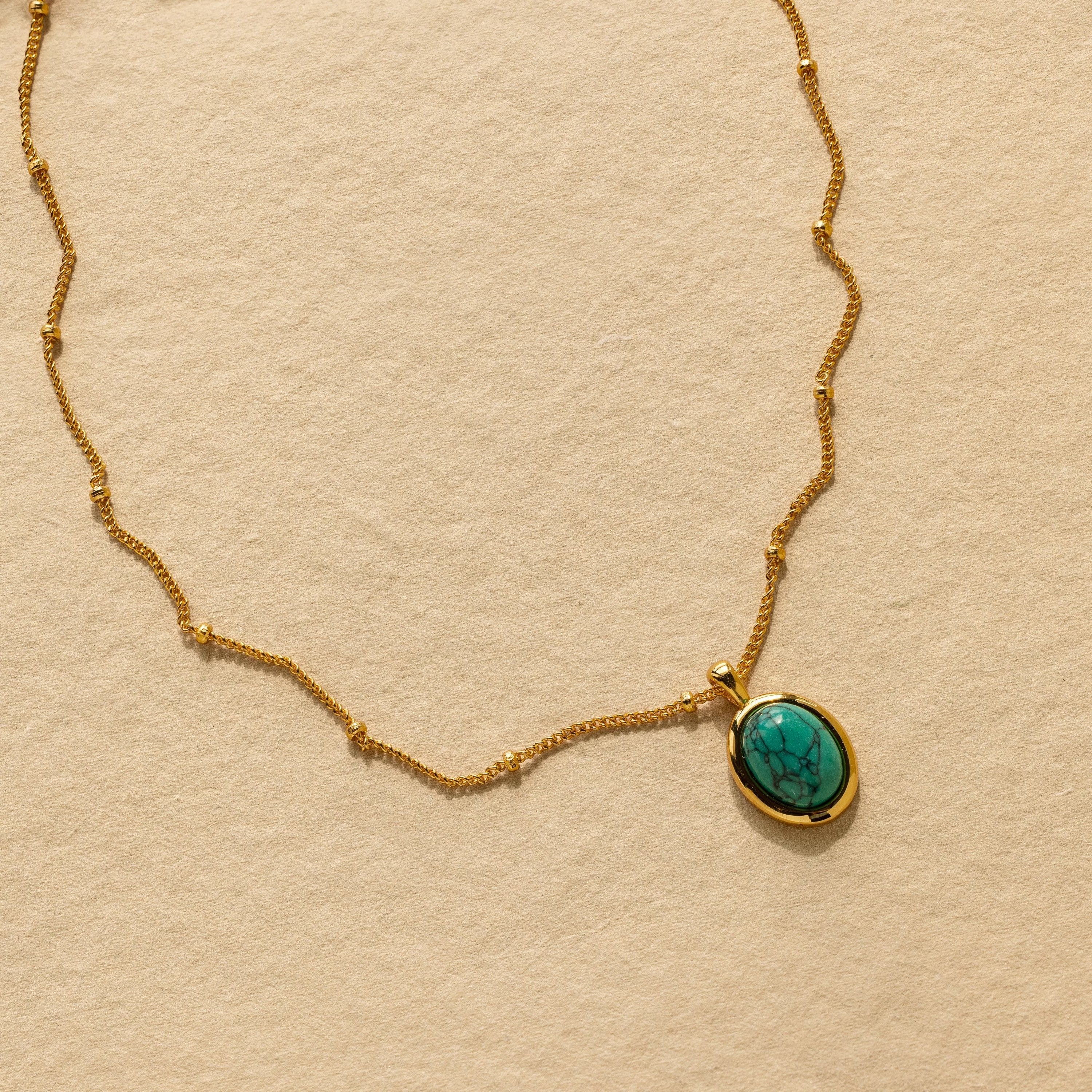 The Turquoise Pendant Necklace features a gold chain and an oval turquoise pendant, styled on a beige textured background for a subtle boho-chic vibe.