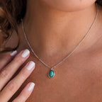 A woman wearing the Turquoise Pendant Necklace—a boho-chic silver chain with an oval turquoise pendant—gently touches her collarbone.