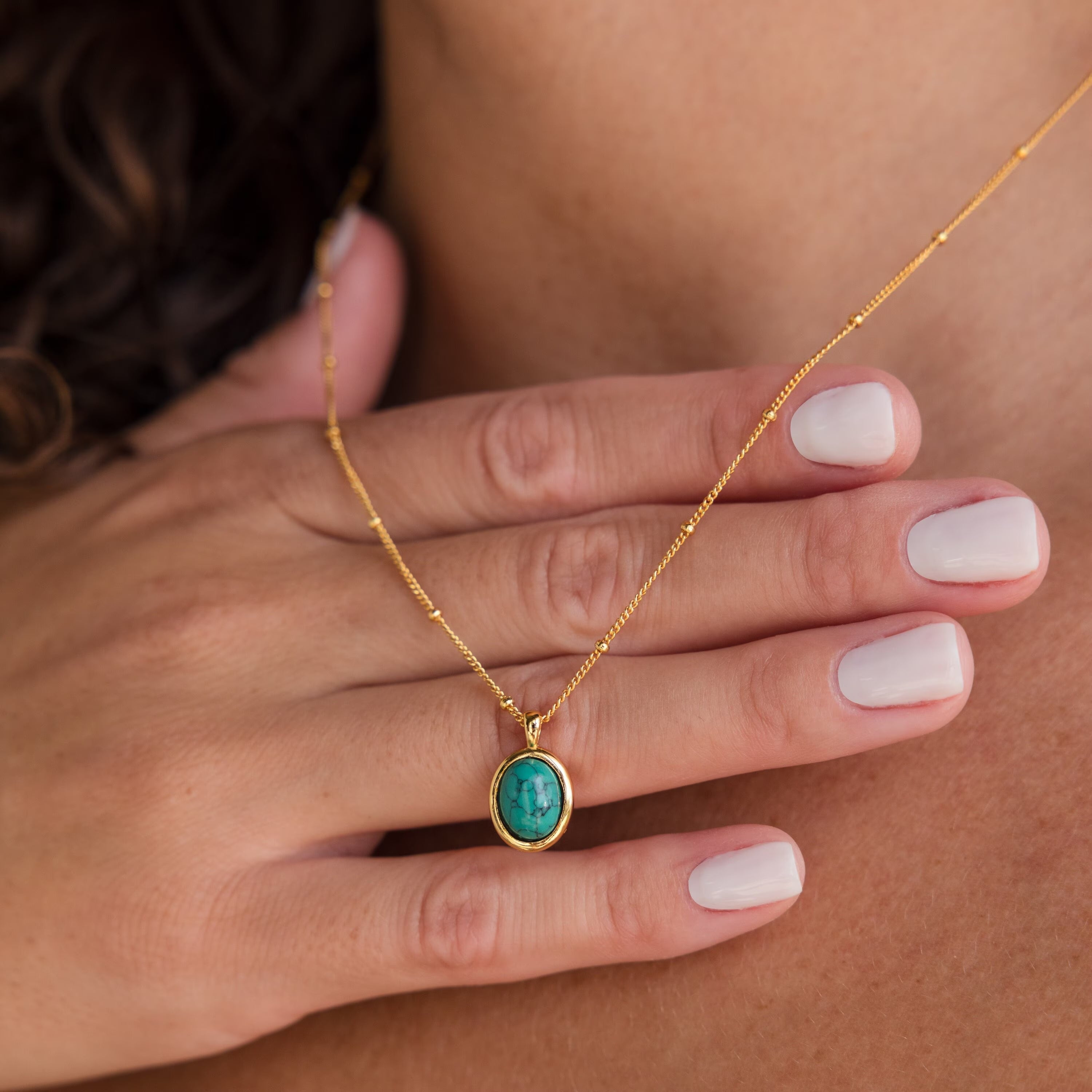 A hand with pale nails touches the Turquoise Pendant Necklace, a boho-chic piece featuring an oval turquoise pendant adorning a person's neck.