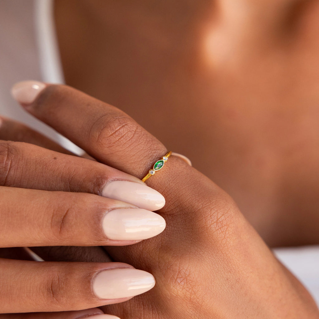 A hand with light pink nails showcases the Marquise Bezel Birthstone Ring, a delicate, vintage-inspired gold ring with a small green gemstone—ideal as a customizable birthstone piece for a personal touch.