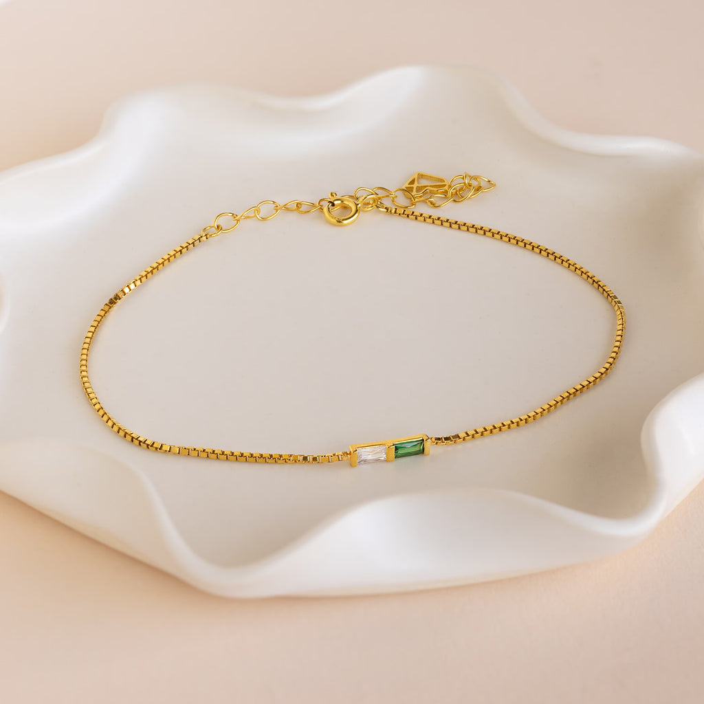 The Multiple Baguette Birthstone Bracelet features a gold box chain with small rectangular green and clear stones, elegantly displayed on a white wavy ceramic dish—perfect for any occasion.