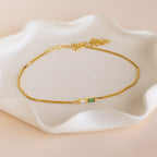The Multiple Baguette Birthstone Bracelet features a gold box chain with small rectangular green and clear stones, elegantly displayed on a white wavy ceramic dish—perfect for any occasion.