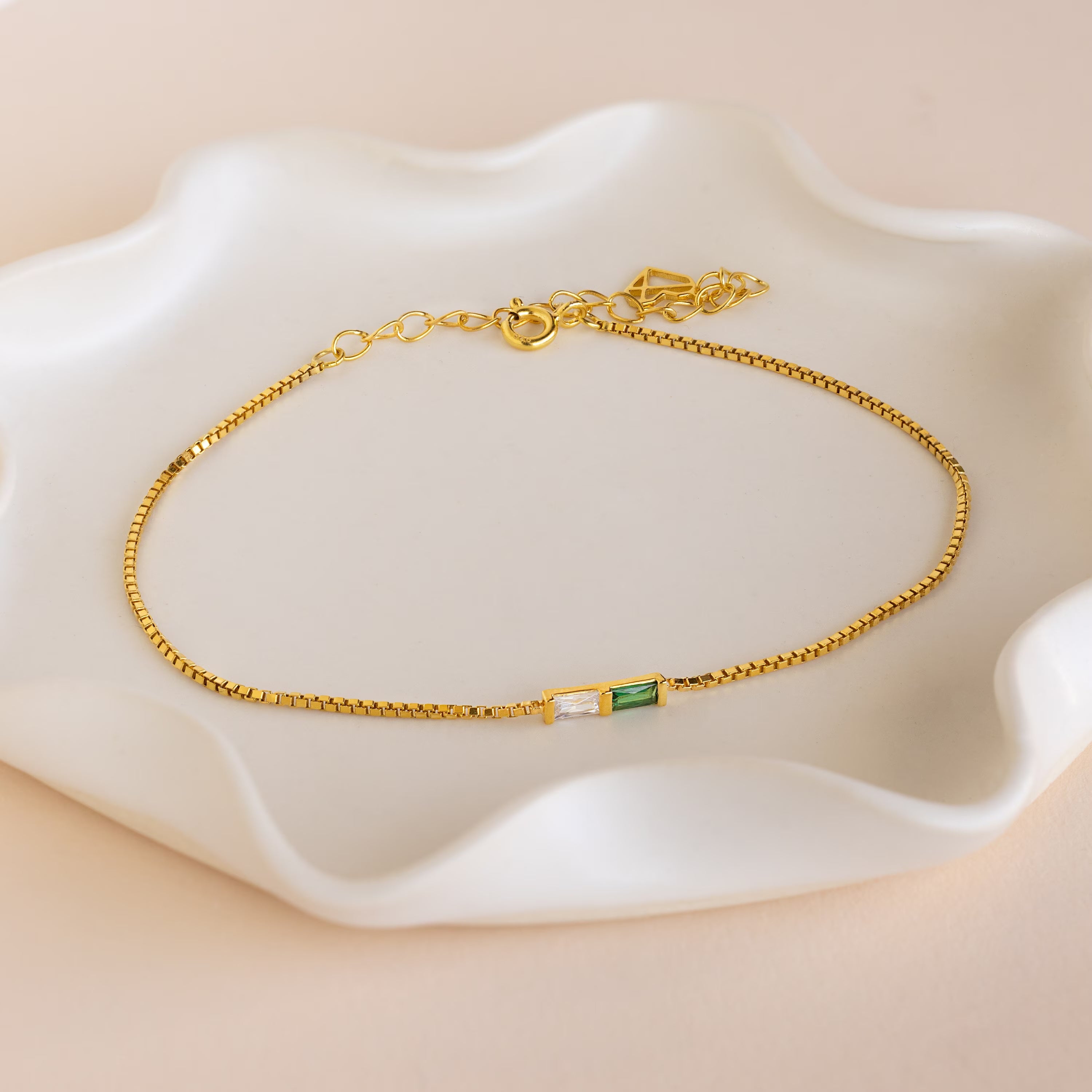The Multiple Baguette Birthstone Bracelet features a gold box chain with small rectangular green and clear stones, elegantly displayed on a white wavy ceramic dish—perfect for any occasion.