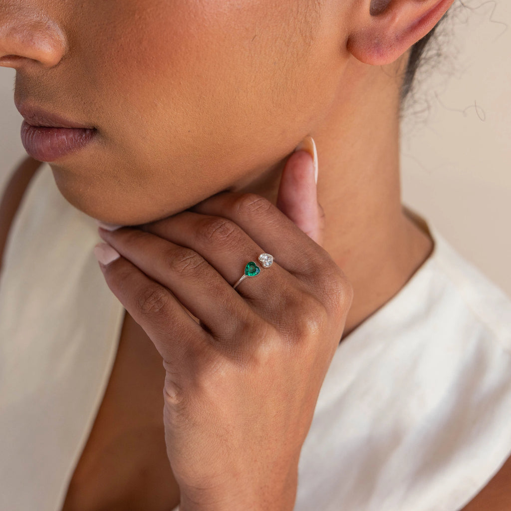 A woman touches her neck while wearing the Duo Heart Birthstone Ring, featuring green and clear heart-shaped gemstones on her finger.