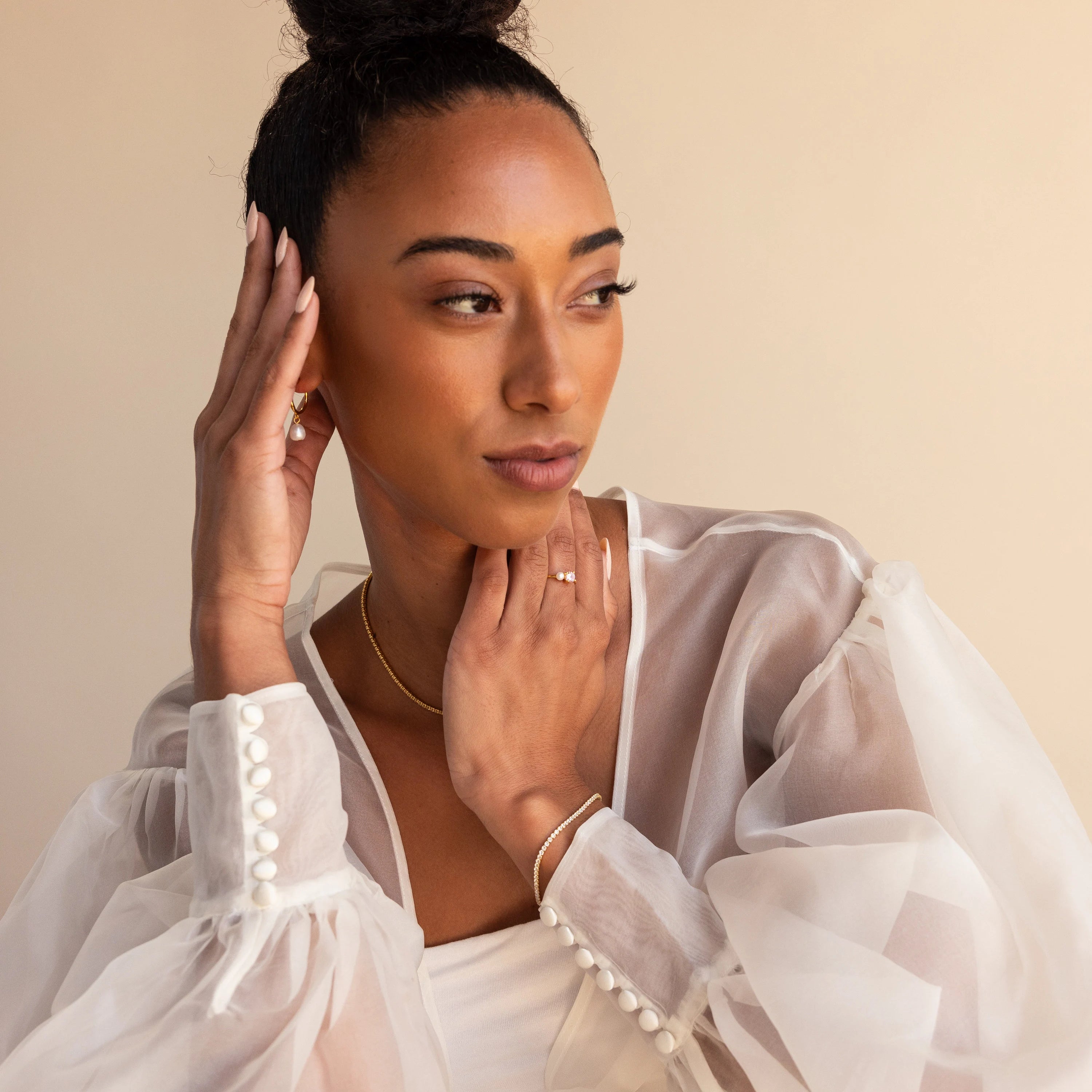 Woman wearing a sheer white blouse and gold jewelry, including the Birthstone & Pearl Ring, poses with her hand on her face against a neutral backdrop.