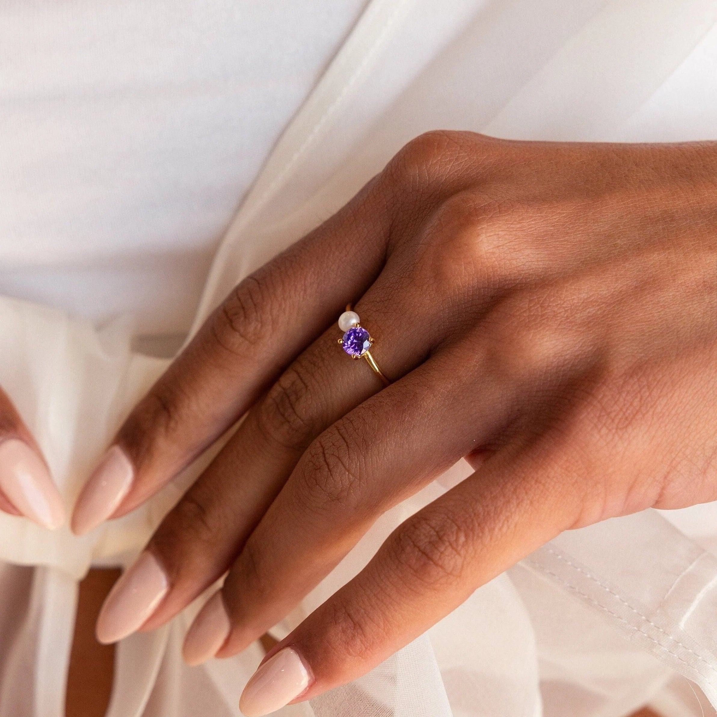 A nude-polished hand wears the Birthstone & Pearl Ring, a gold ring with a purple stone and pearl accent.