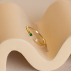 The Duo Birthstone & Opal Ring, featuring green and white stones, rests on a wavy beige surface, blending unique style with classic elegance.