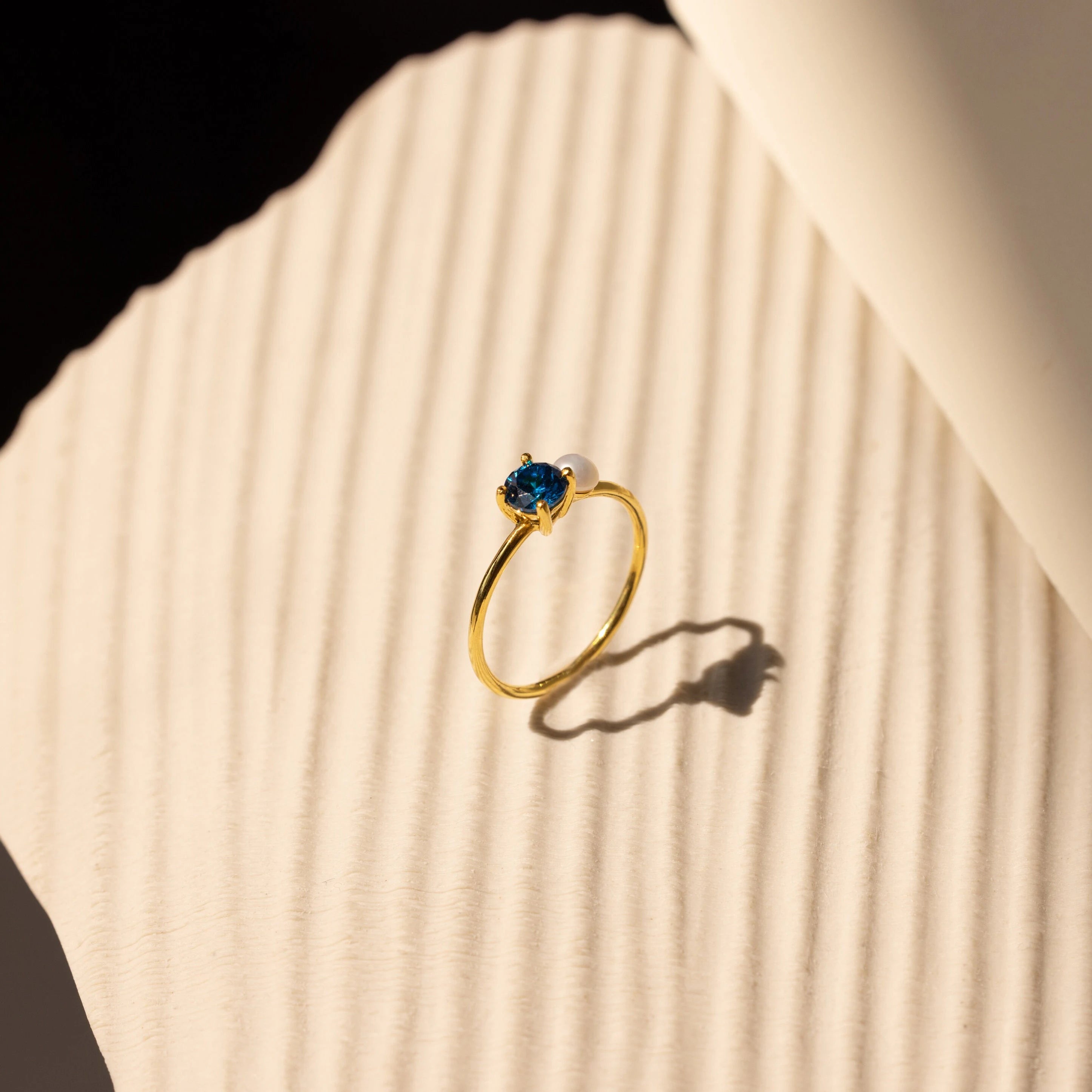 The Birthstone & Pearl Ring features a blue gemstone and a delicate pearl, displayed on a textured cream surface in soft sunlight—ideal for lovers of personalized jewelry.