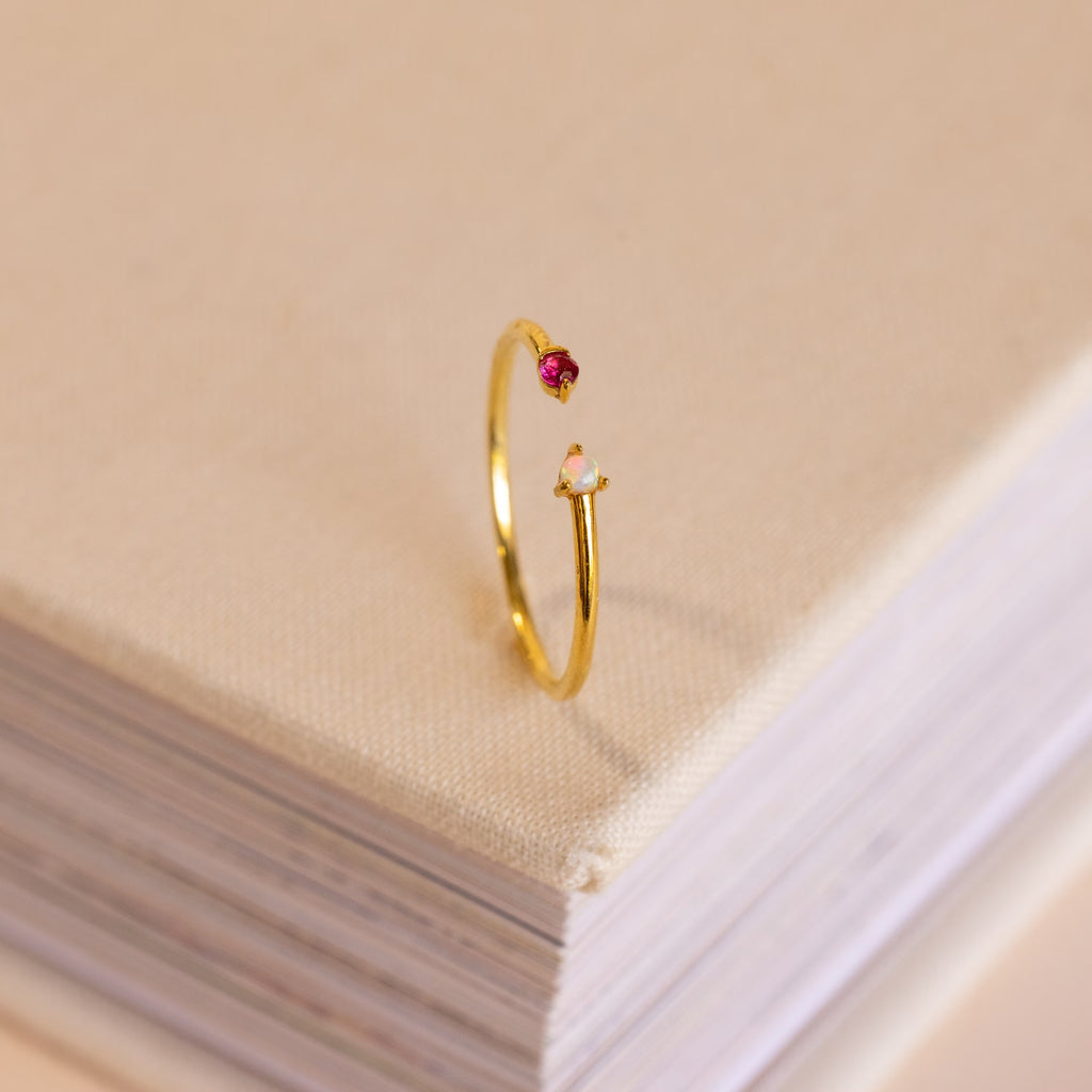 The Duo Birthstone & Opal Ring, featuring two small gemstones, rests on the corner of a closed, fabric-covered book.