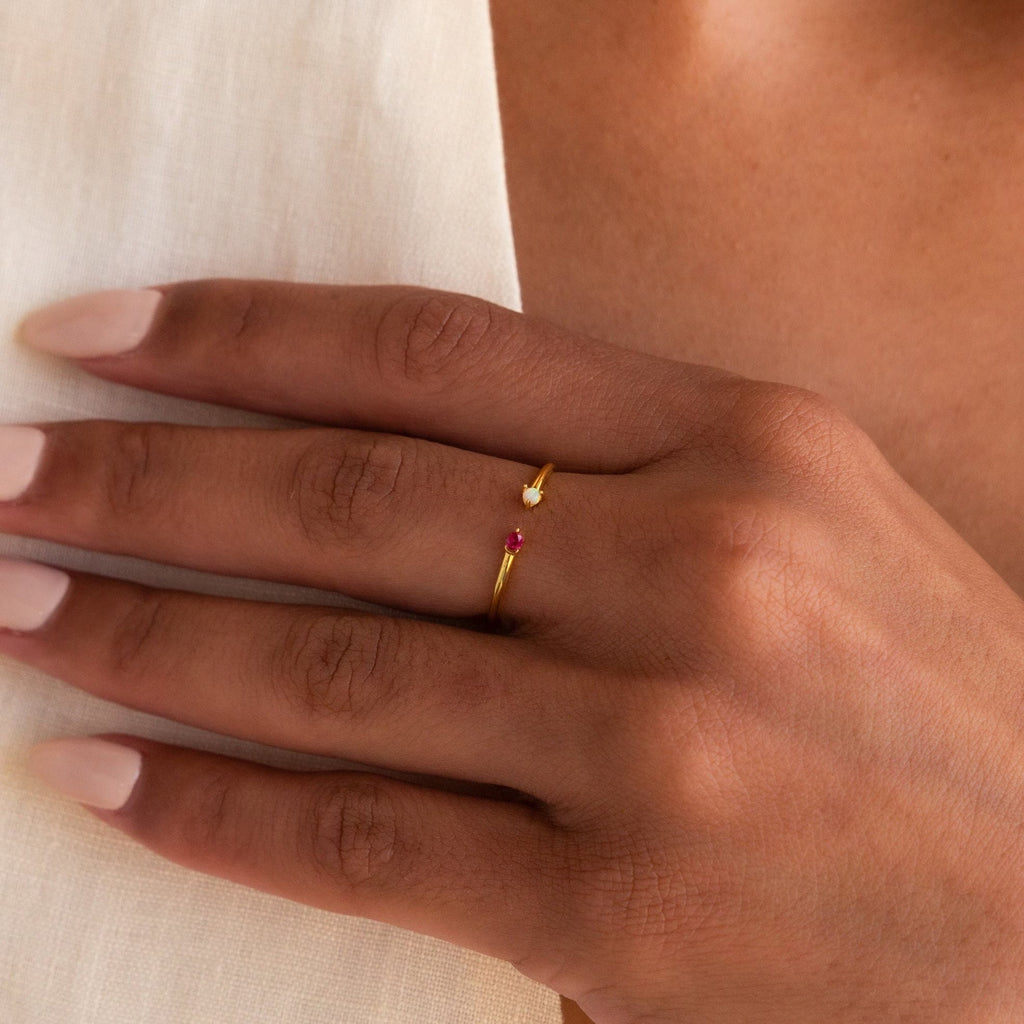 A hand with nude nails wears the Duo Birthstone & Opal Ring, a delicate gold customizable ring featuring a small red stone on the ring finger.