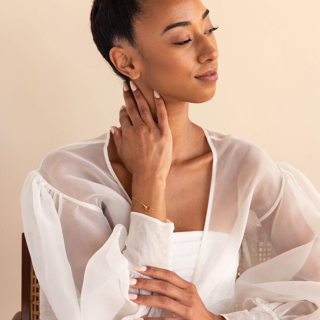 Woman in a sheer white blouse touches her neck while wearing the Interlocking Birthstone Bracelet, posed against a beige background.