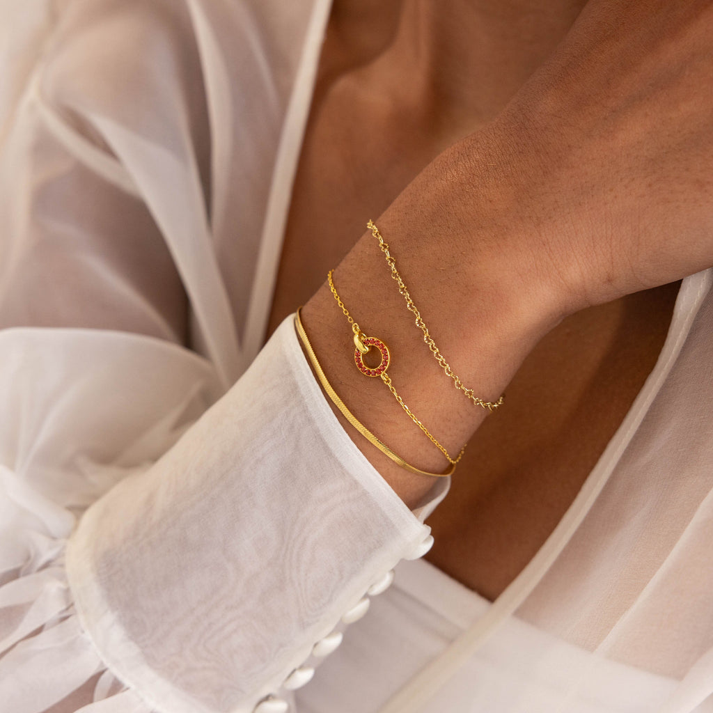 A woman wears gold bracelets, including the Interlocking Birthstone Bracelet, a beautiful customizable piece, and a sheer white blouse with buttoned cuffs.