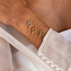 A hand wears three Interlocking Birthstone Bracelets—gold chains with circular gemstone charms—resting on white fabric. This stunning birthstone jewelry is perfect for stacking or gifting.