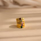 The Ribbed Birthstone Signet Ring is stacked with four gold bands featuring colorful gemstones, all displayed on a beige surface in soft, natural light.