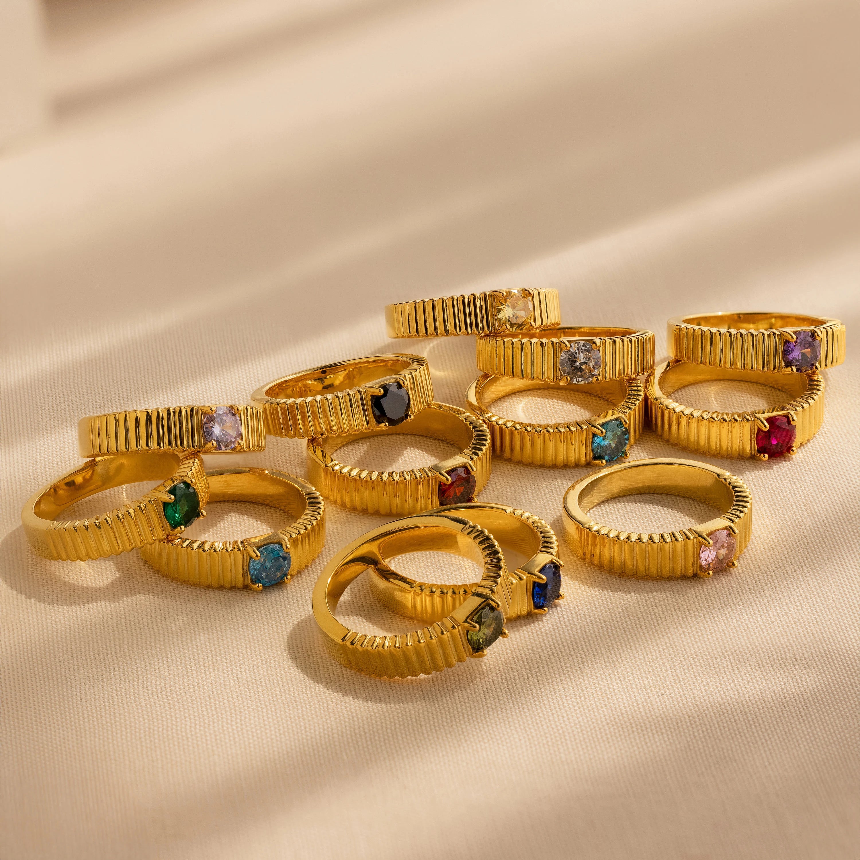 Twelve Ribbed Birthstone Signet Rings, each featuring a unique colored gemstone, are displayed on a beige surface—perfect for personalizing as a birthstone or signet ring.