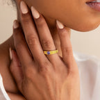 A hand with nude nails wears the Ribbed Birthstone Signet Ring, featuring a gold band with ribbed detailing and a purple gemstone, as it touches the side of the face.
