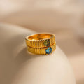 The Ribbed Birthstone Signet Ring features a gold ribbed band set with a vibrant gemstone, creating a modern look against a soft beige background.