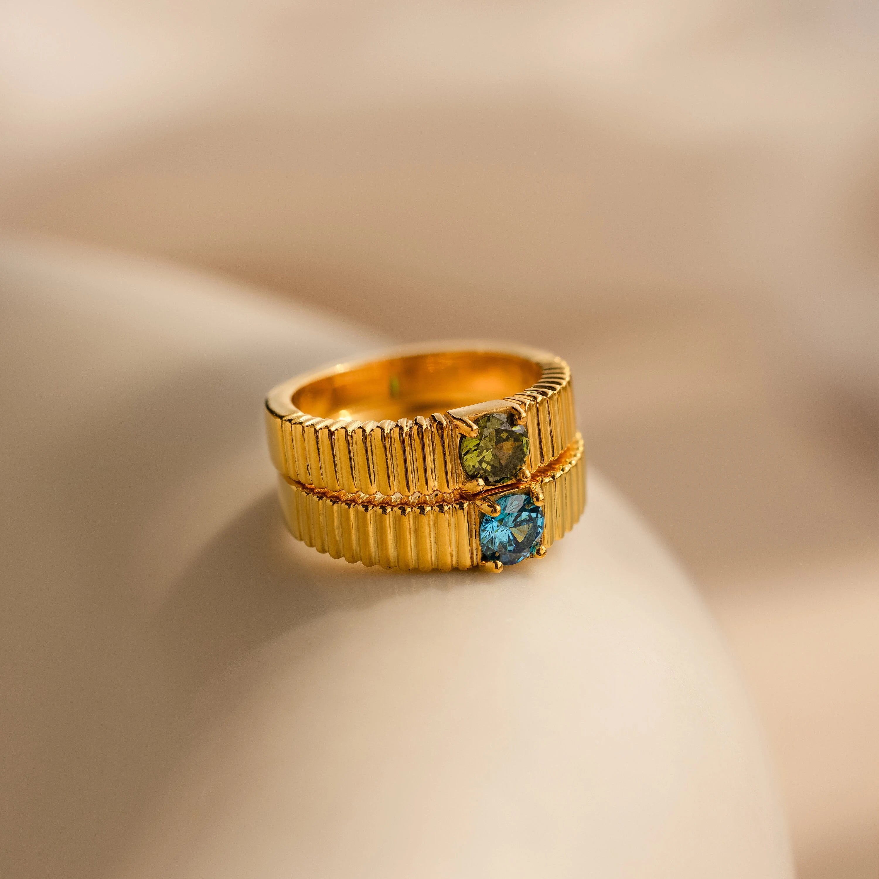 The Ribbed Birthstone Signet Ring features a gold ribbed band set with a vibrant gemstone, creating a modern look against a soft beige background.
