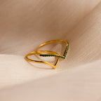 The Chevron Birthstone Rings Set features a gold ring with a V-shaped chevron design, accented with small green stones, displayed on light beige fabric. Ideal for anyone searching for distinctive Chevron Birthstone Rings.