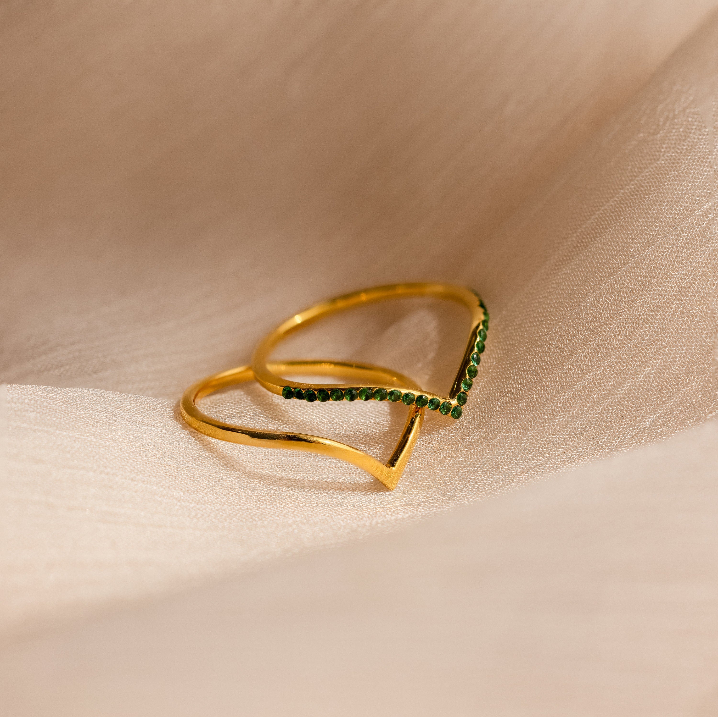 The Chevron Birthstone Rings Set features a gold ring with a V-shaped chevron design, accented with small green stones, displayed on light beige fabric. Ideal for anyone searching for distinctive Chevron Birthstone Rings.