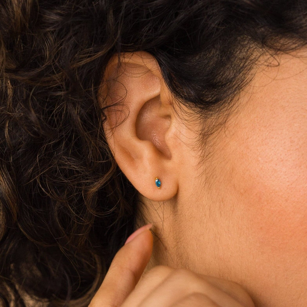 Close-up of a person’s ear with curly hair, wearing Marquise Birthstone Studs featuring a turquoise gemstone.